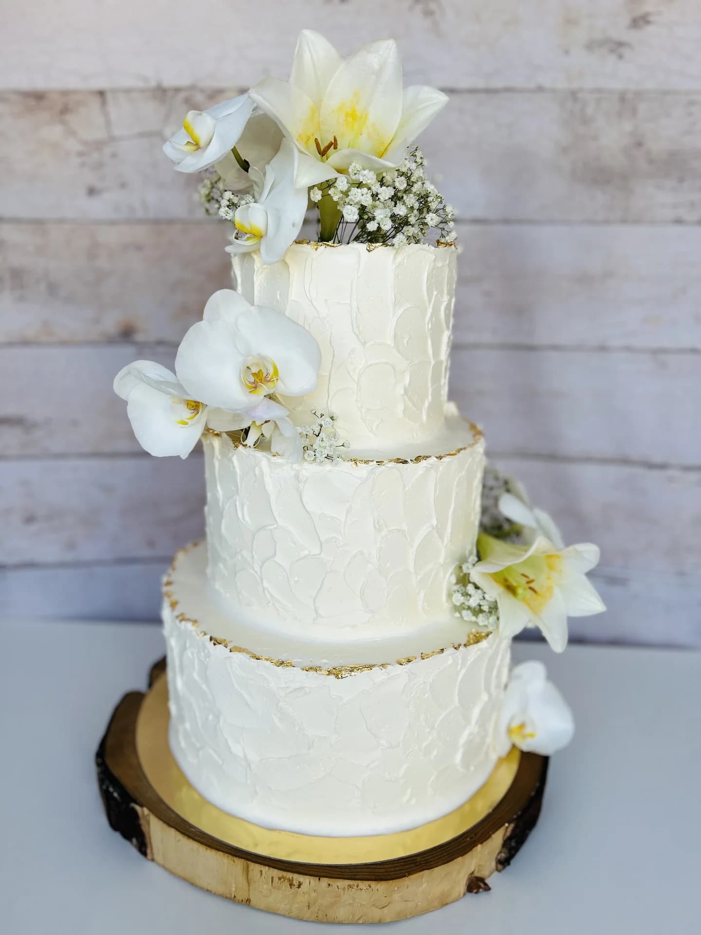 Dreistöckige weiße Hochzeitstorte mit Orchideen, Lilien und Goldblatt-Akzenten